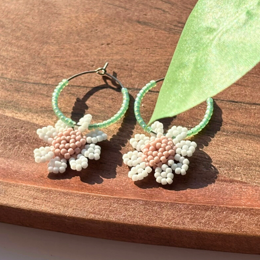 Cercei hoops handmade cu cerc verde mentă din mărgele și floricele albe cu centru roz pudrat, fotografiați pe suport din lemn – accesorii florale delicate, perfecte pentru primăvară/vară și cadou.