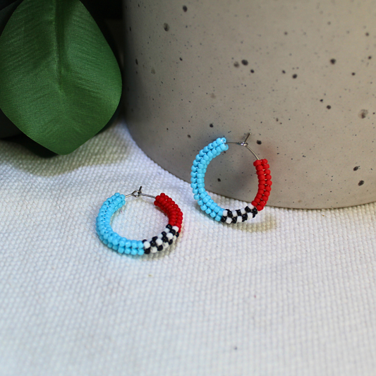 Cercei tip hoop realizați manual din mărgele turcoaz, roșii, alb și negru, cu design modern și contrastant, fotografiați pe fundal textil natural cu accente botanice – bijuterii handmade ușoare, ideale pentru ținute casual sau cadou.