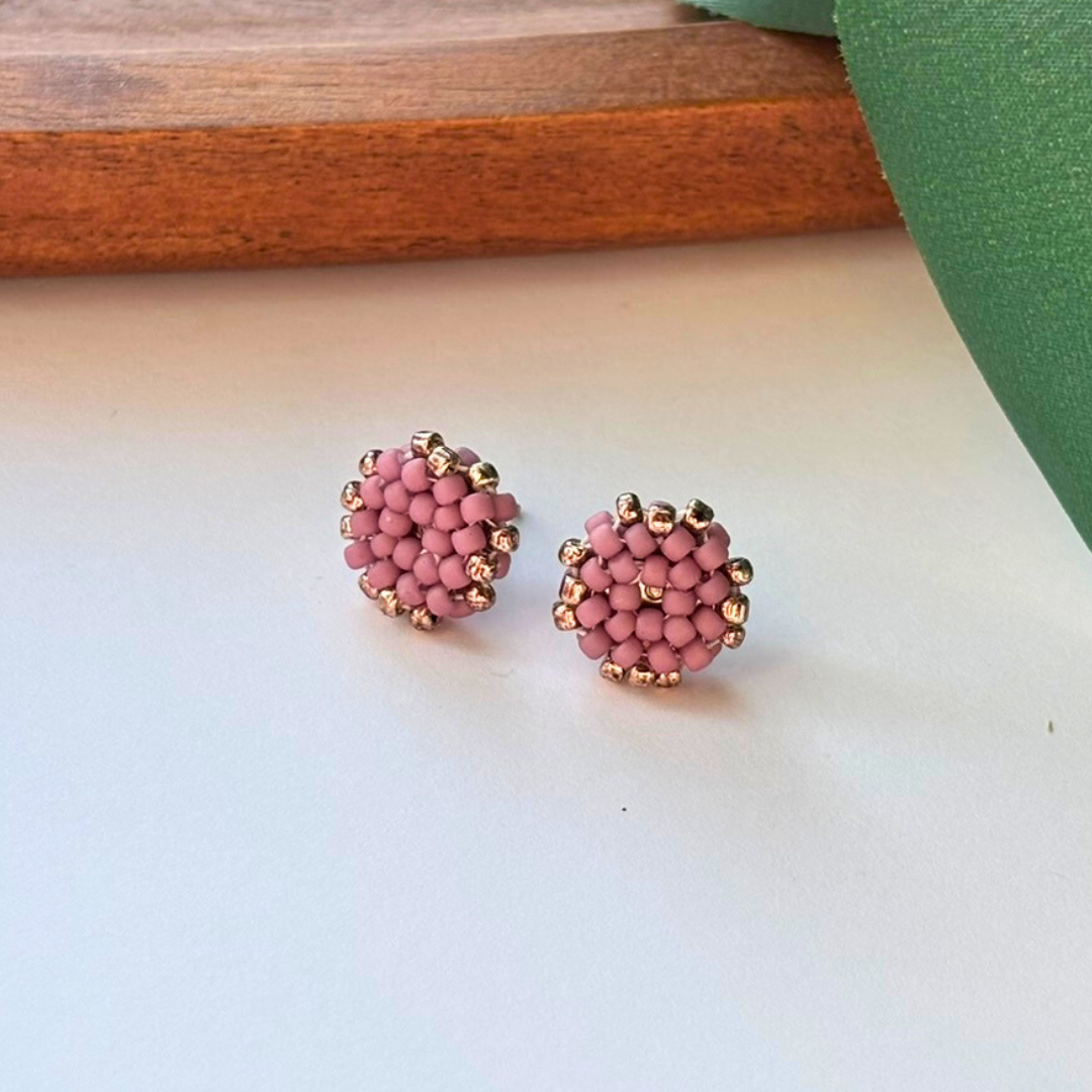 Cercei handmade tip stud, rotunzi, lucrati manual, din mărgele roz prăfuit cu accente aurii, fotografiați pe fundal natural – bijuterii minimaliste, feminine, ideale pentru ținute elegante sau cadou.