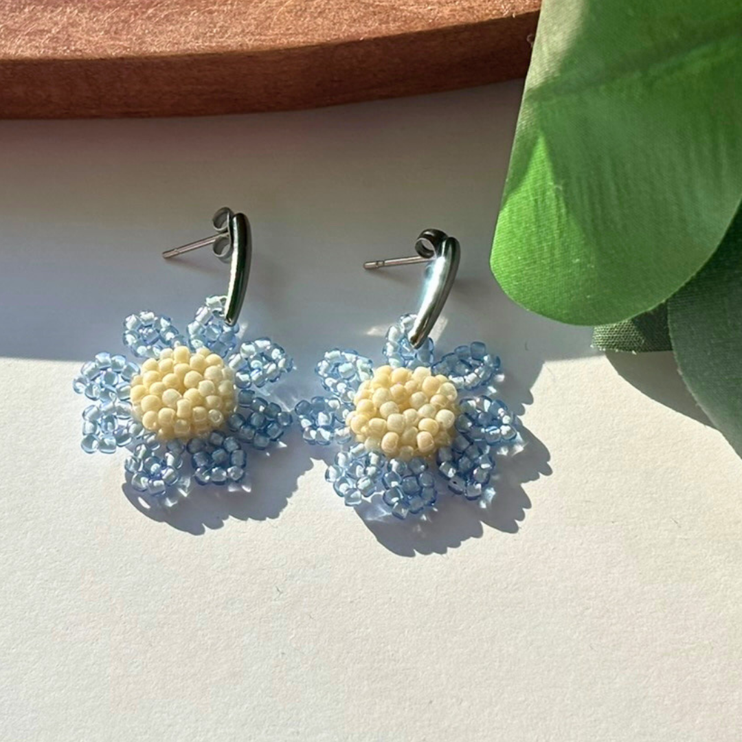 Cercei handmade tip floare/margaretă, realizați din mărgele albastre transparente cu centru crem, cu prindere metalică – accesorii florale ușoare, ideale pentru primăvară și vară.