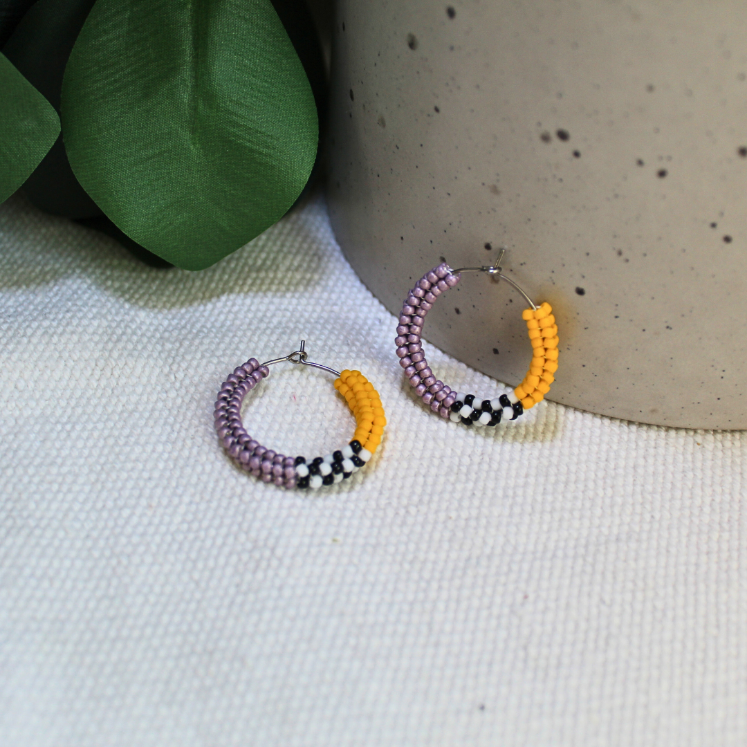 Cercei tip hoop realizați manual din mărgele mov și galben muștar, cu detalii alb-negru și tortițe metalice, fotografiați pe fundal textil natural cu elemente botanice – bijuterii handmade moderne, ușoare, ideale pentru ținute casual sau cadou.