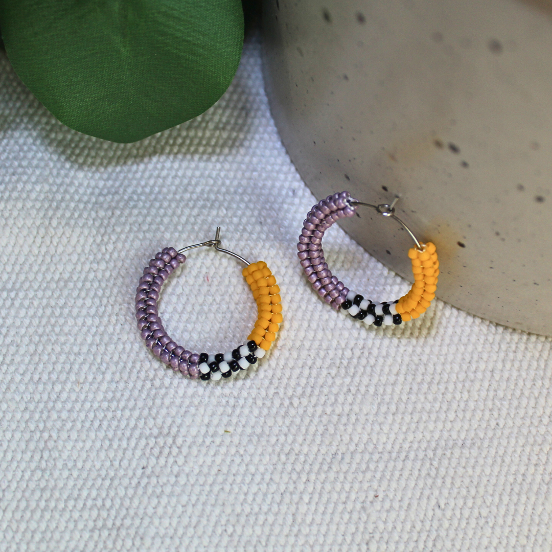 Cercei tip hoop realizați manual din mărgele mov și galben muștar, cu detalii alb-negru și tortițe metalice, fotografiați pe fundal textil natural cu elemente botanice – cercei handmade moderni, ușori, perfecți pentru ținute casual sau cadou special.