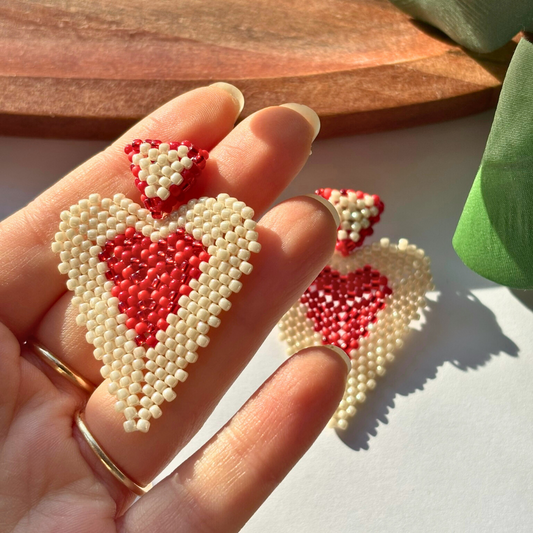 Cercei handmade lucrați manual în formă de inimă, din mărgele crem cu interior roșu, fotografiați în mână pentru detalii – bijuterii statement romantice, ideale pentru cadou de Valentine’s Day sau aniversare.