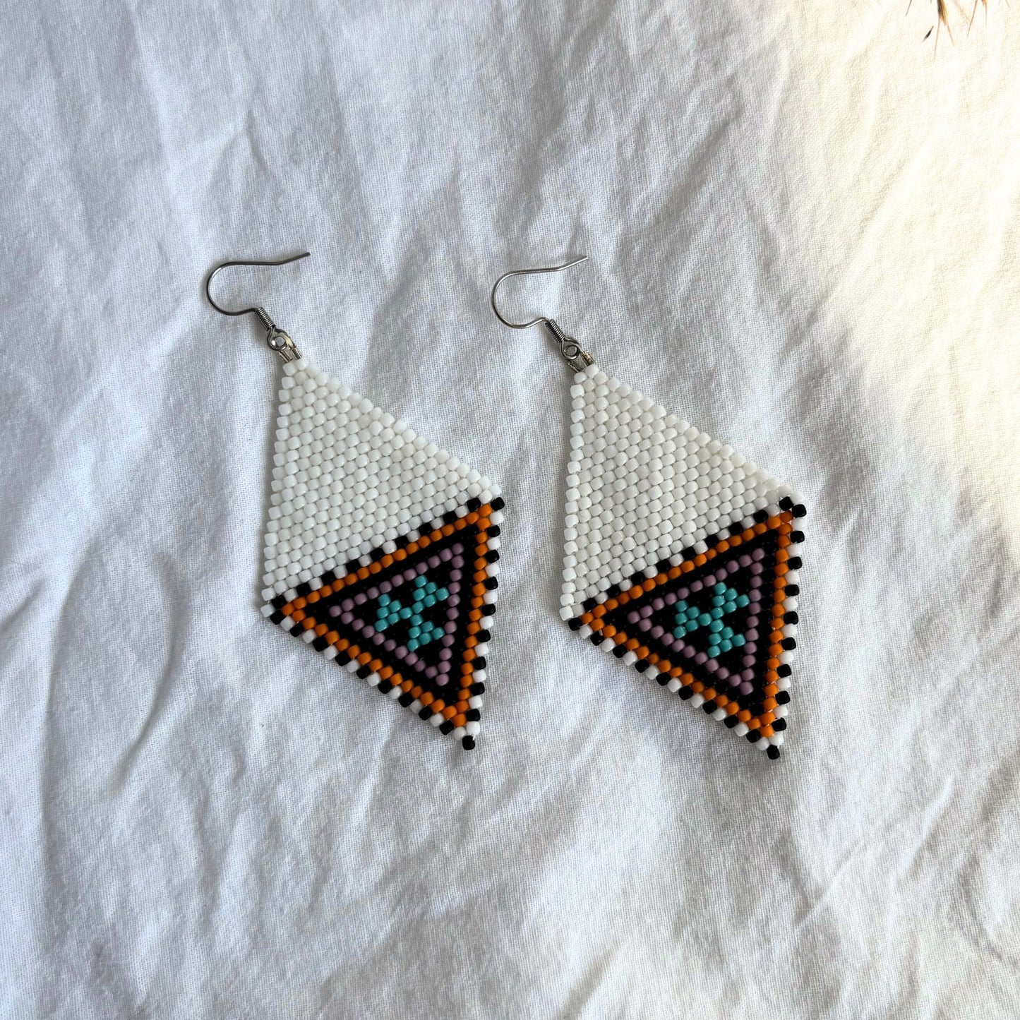 Cercei handmade lungi din mărgele delicate în nuanțe de alb, negru, portocaliu, mov și accente turcoaz, design geometric tip romb, cercei statement ușori, stil boho elegant și modern, bijuterii artizanale unicat potrivite pentru ținute de zi sau de ocazie.