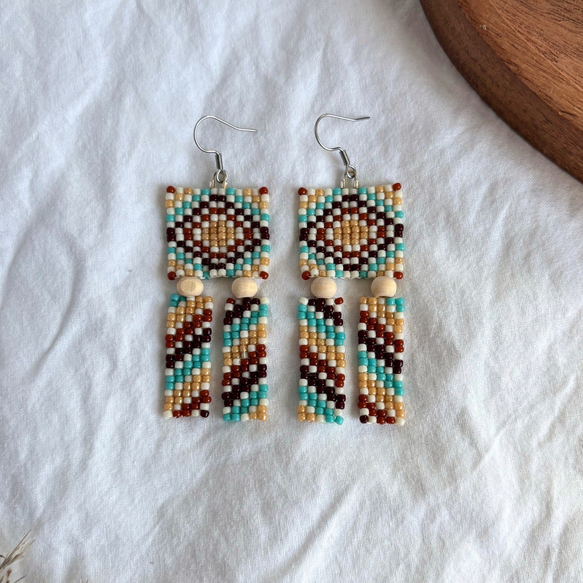 Cercei lungi handmade din mărgele, model geometric multicolor cu nuanțe de turcoaz, bej, maro și alb, accente din lemn natural, bijuterii artizanale boho, ușori și expresivi, potriviți pentru ținute casual sau de vară.