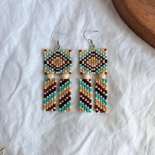 Cercei lungi handmade din mărgele, model geometric multicolor cu nuanțe de turcoaz, bej, maro și alb, accente din lemn natural, bijuterii artizanale boho, ușori și expresivi, potriviți pentru ținute casual sau de vară.