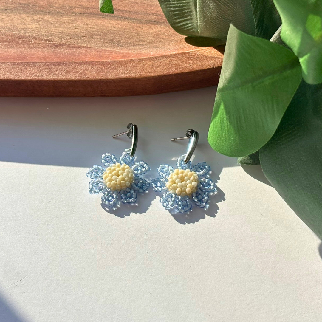 Cercei handmade în formă de margaretă, din mărgele albastre transparente cu centru crem, pe tortițe metalice – bijuterii florale delicate, perfecte pentru ținute de primăvară/vară.