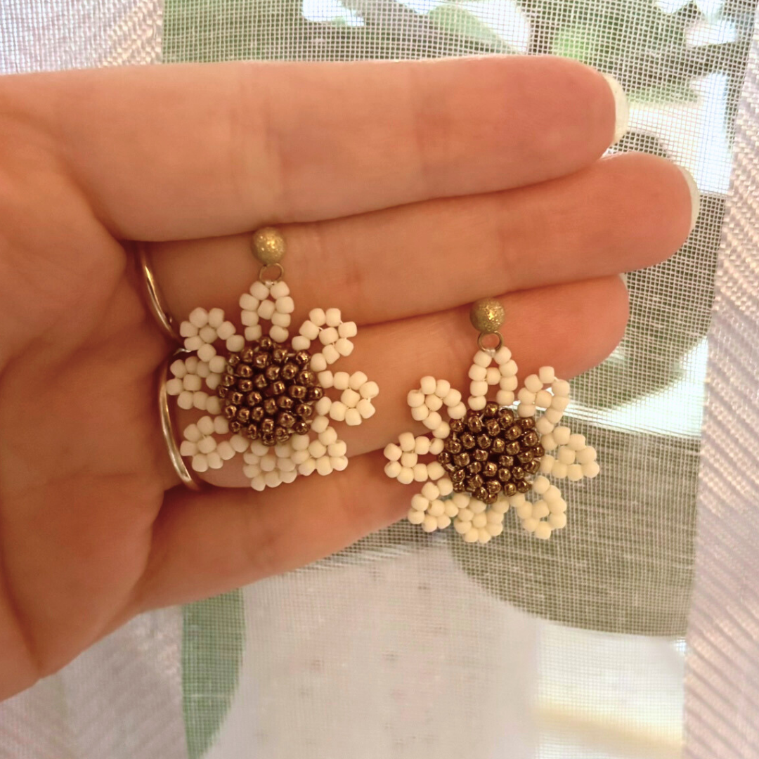 Cercei handmade tip margaretă, din mărgele albe cu centru bronz și prindere aurie tip șurub, fotografiați în mână – bijuterii florale delicate, ideale de purtat zilnic sau cadou.