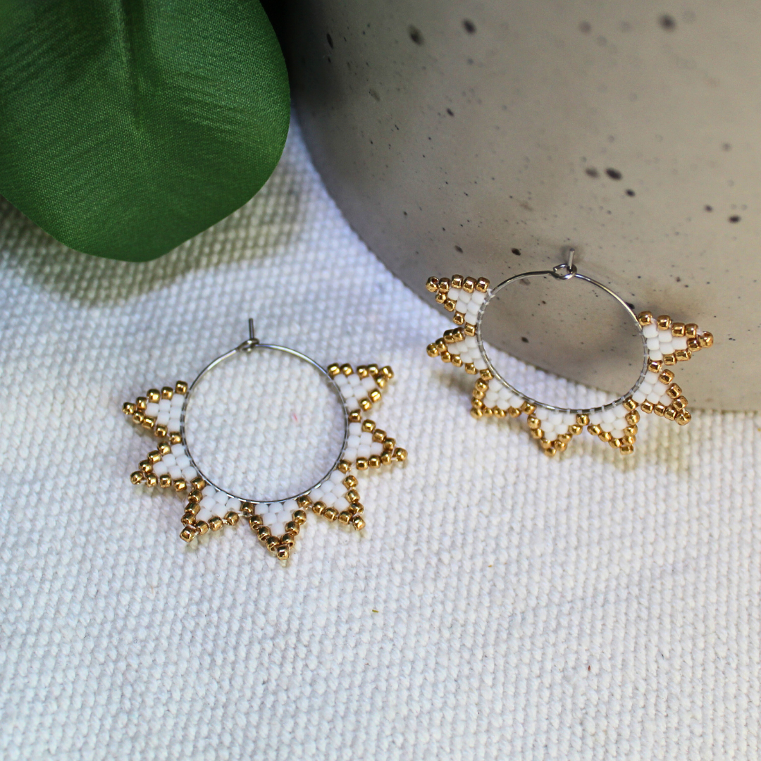 Cercei handmade tip hoop în formă de soare, realizați din mărgele albe cu accente aurii, design delicat cu raze stilizate, fotografiați pe fundal textil natural cu elemente botanice – cercei statement ușori, ideali pentru ținute elegante, de vară sau cadou special.
