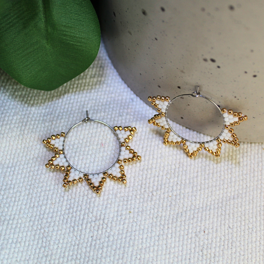 Cercei handmade tip hoop în formă de soare, realizați din mărgele albe cu accente aurii, design delicat cu raze stilizate, fotografiați pe fundal textil natural cu elemente botanice – cercei statement ușori, ideali pentru ținute elegante, de vară sau cadou special.