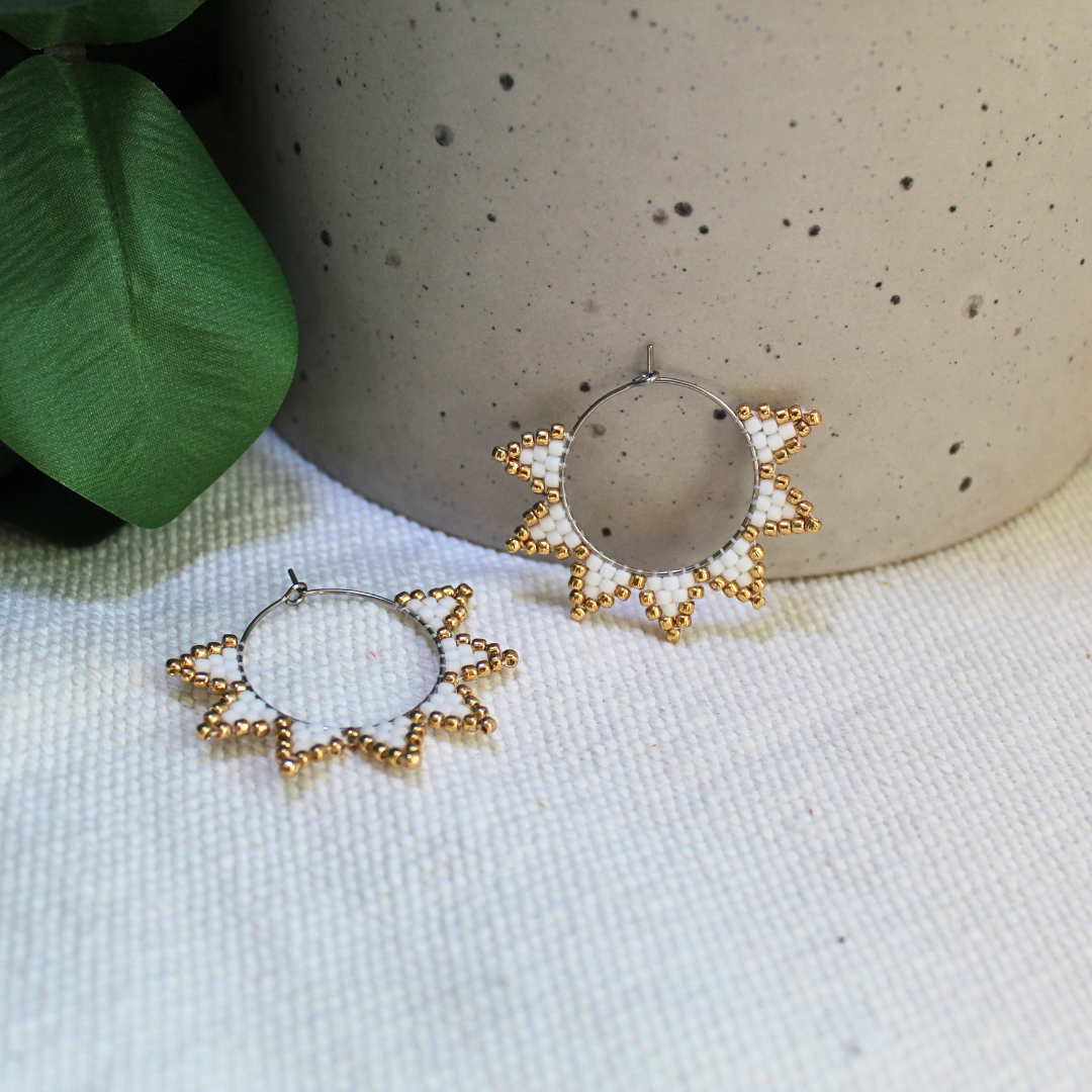 Cercei handmade tip hoop în formă de soare, realizați din mărgele albe cu accente aurii, design geometric inspirat de raze solare, fotografiați pe fundal textil natural cu elemente botanice – cercei statement eleganți, ușori, perfecți pentru ținute de vară sau cadou special.
