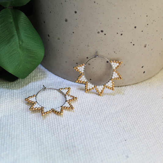 Cercei handmade tip hoop în formă de soare, realizați din mărgele albe cu accente aurii, design geometric inspirat de raze solare, fotografiați pe fundal textil natural cu elemente botanice – cercei statement eleganți, ușori, perfecți pentru ținute de vară sau cadou special.