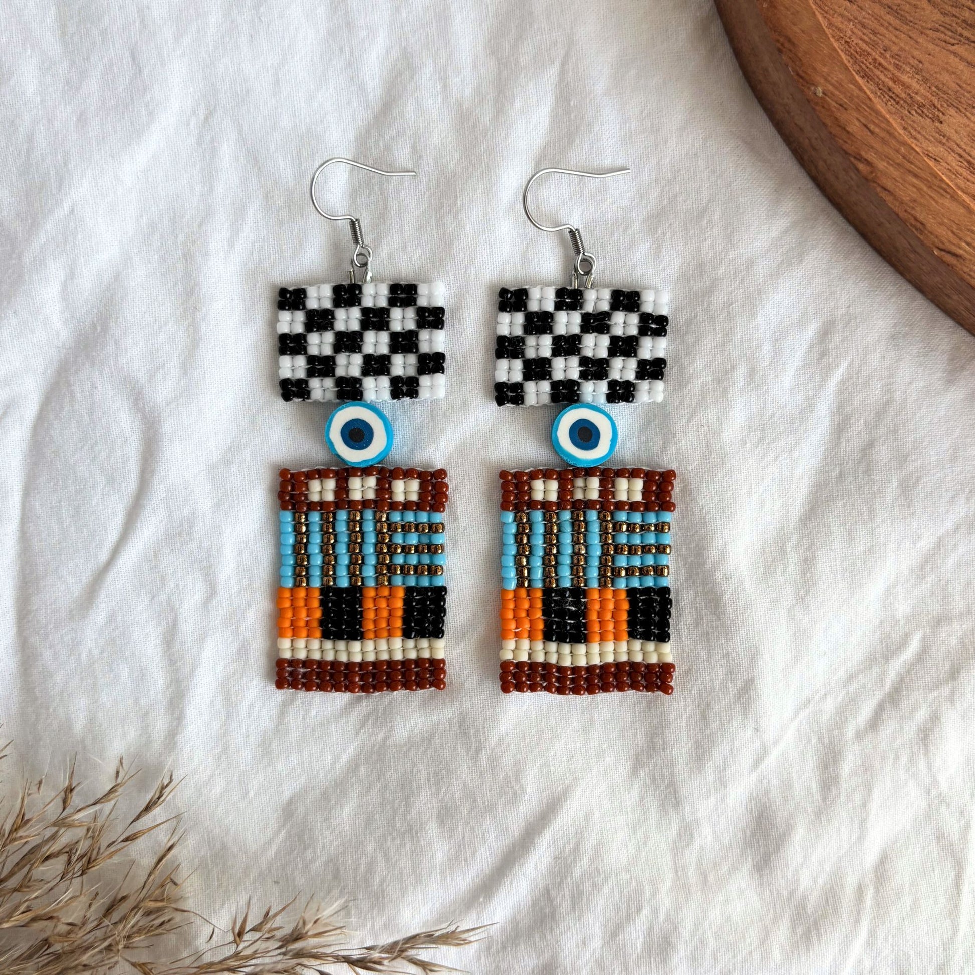 Cercei handmade din mărgele fine, design geometric urban cu detaliu ochi protector, combinație de alb, negru, turcoaz, portocaliu și maro, cercei statement etnici moderni, bijuterii artizanale unicat, stil boho contemporan, accesorii originale pentru ținute creative.