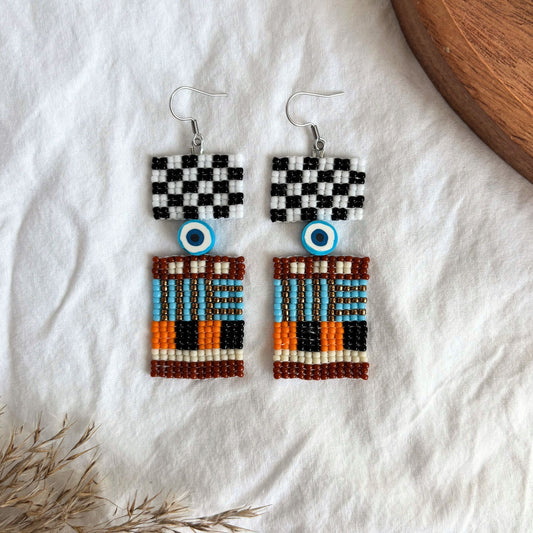 Cercei handmade din mărgele fine, design geometric urban cu detaliu ochi protector, combinație de alb, negru, turcoaz, portocaliu și maro, cercei statement etnici moderni, bijuterii artizanale unicat, stil boho contemporan, accesorii originale pentru ținute creative.