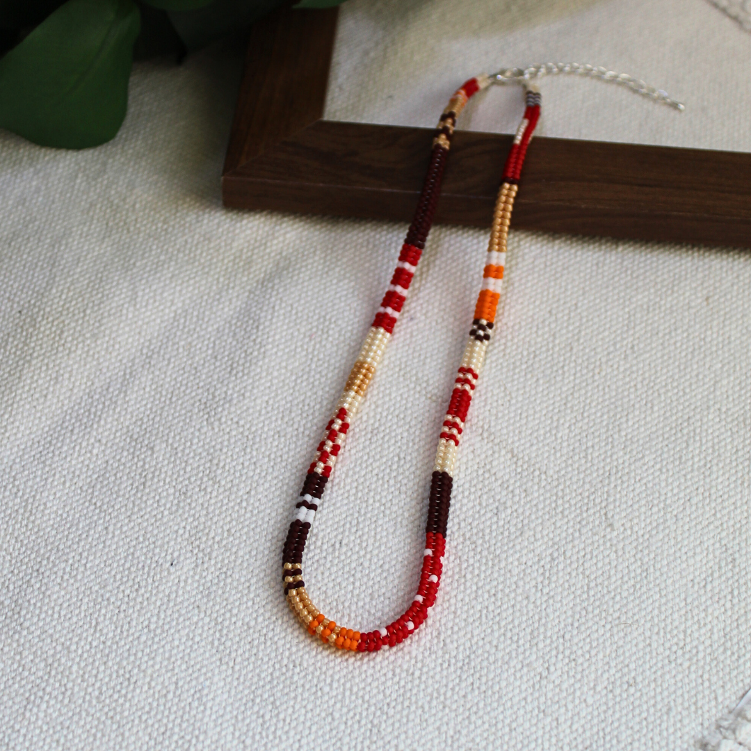 Colier handmade lucrat manual din mărgele fine în nuanțe de roșu, portocaliu, maro și crem, design boho-etnic, așezat pe suport din lemn – bijuterie ușoară, ideală pentru ținute casual, de vară sau cadou special.