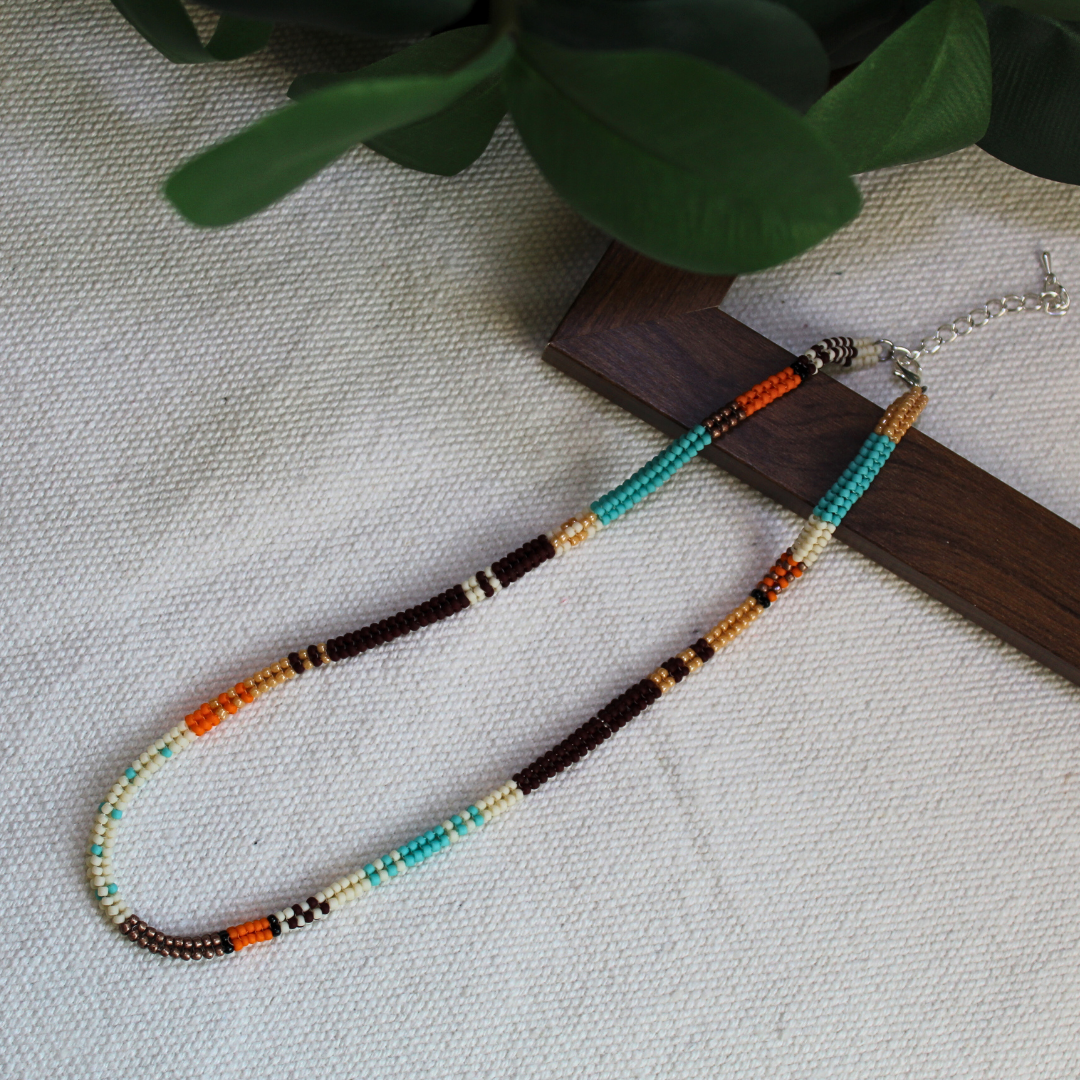 Colier handmade din mărgele fine în nuanțe de turcoaz, maro, crem și portocaliu, design boho-etnic, așezat pe fundal textil cu suport din lemn – bijuterie ușoară, ideală pentru ținute casual, layering sau cadou.