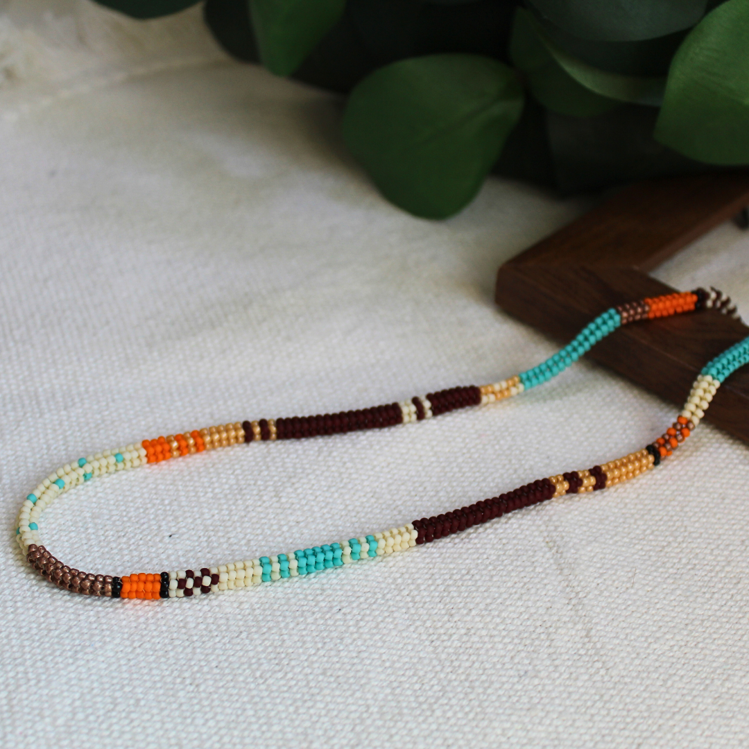 Colier boho realizat manual din mărgele fine în culori turcoaz, maro, crem și portocaliu, cu design etnic minimalist, așezat pe fundal textil natural – bijuterie handmade ușoară, ideală pentru ținute casual sau cadou.