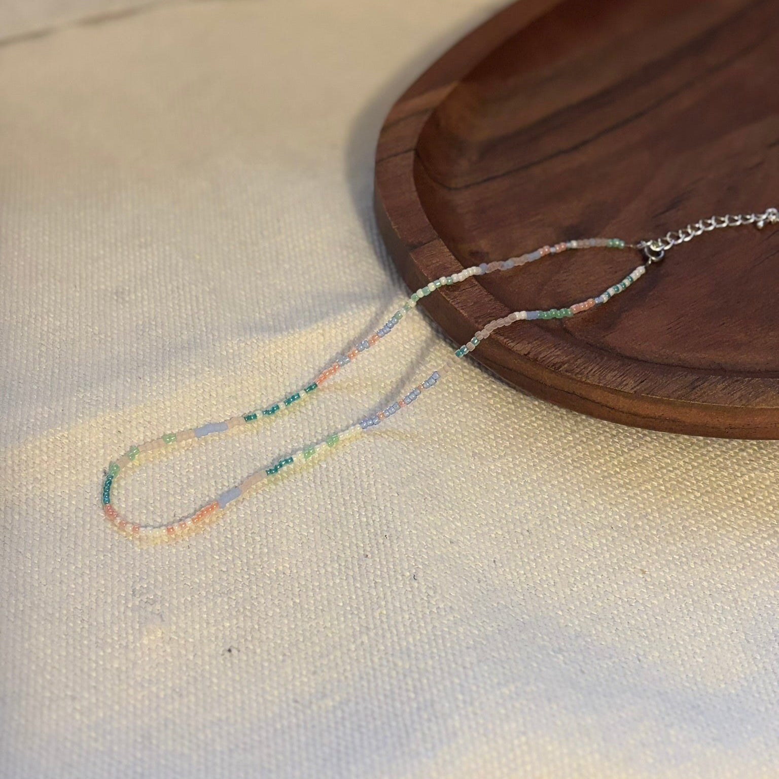 Colier handmade lucrat manual din mărgele fine în nuanțe pastel (roz, albastru, verde și crem), design delicat și minimalist, așezat pe suport din lemn – bijuterie ușoară, ideală pentru purtare zilnică sau cadou.