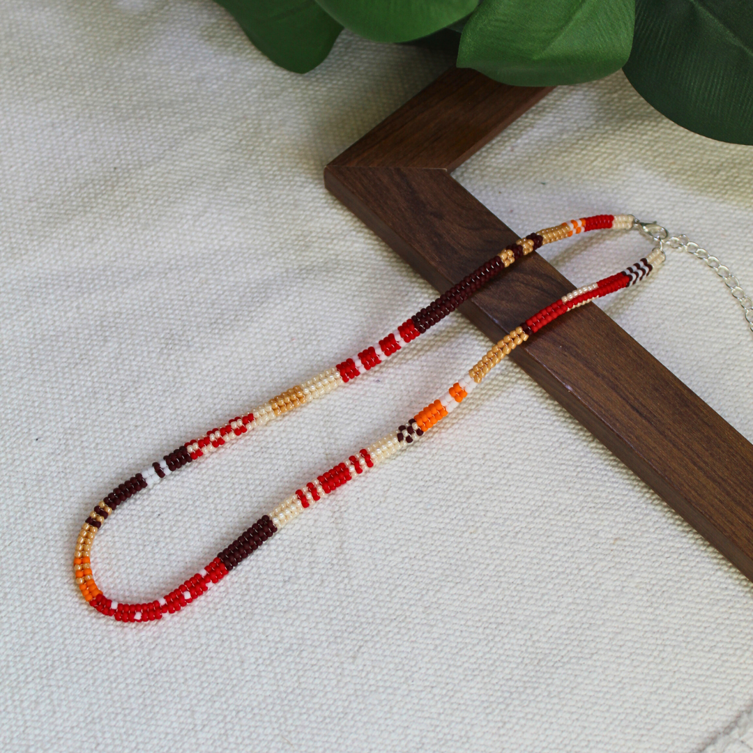 Colier handmade lucrat manual din mărgele fine în nuanțe de roșu, maro, crem și portocaliu, design etnic-boho, așezat pe suport din lemn – bijuterie ușoară, ideală pentru ținute casual, de vară sau cadou special.