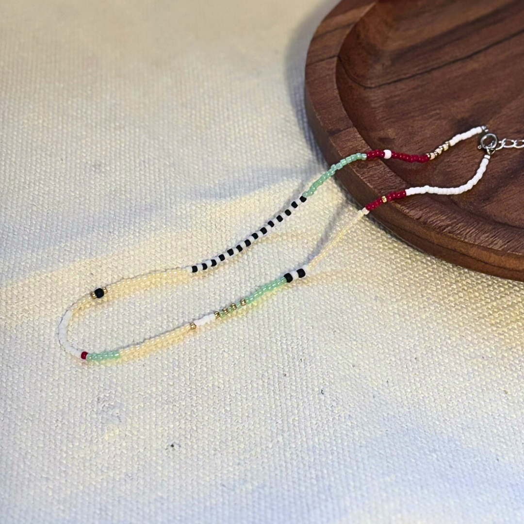 Colier handmade lucrat manual din mărgele fine multicolore (alb, negru, verde mentă, roșu și crem), design minimalist și delicat, așezat pe suport din lemn – bijuterie ușoară, ideală pentru purtare zilnică sau cadou.