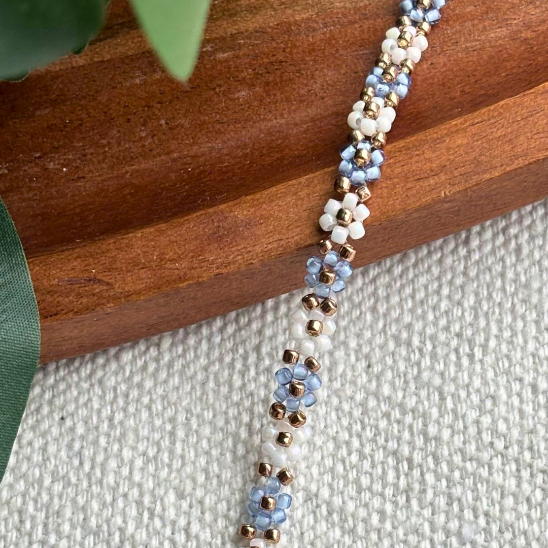 Martisor bratara lucrata manual din margele, cu flori bleu si alb si accente aurii. Cadou elegant pentru ea, perfect pentru primavara.