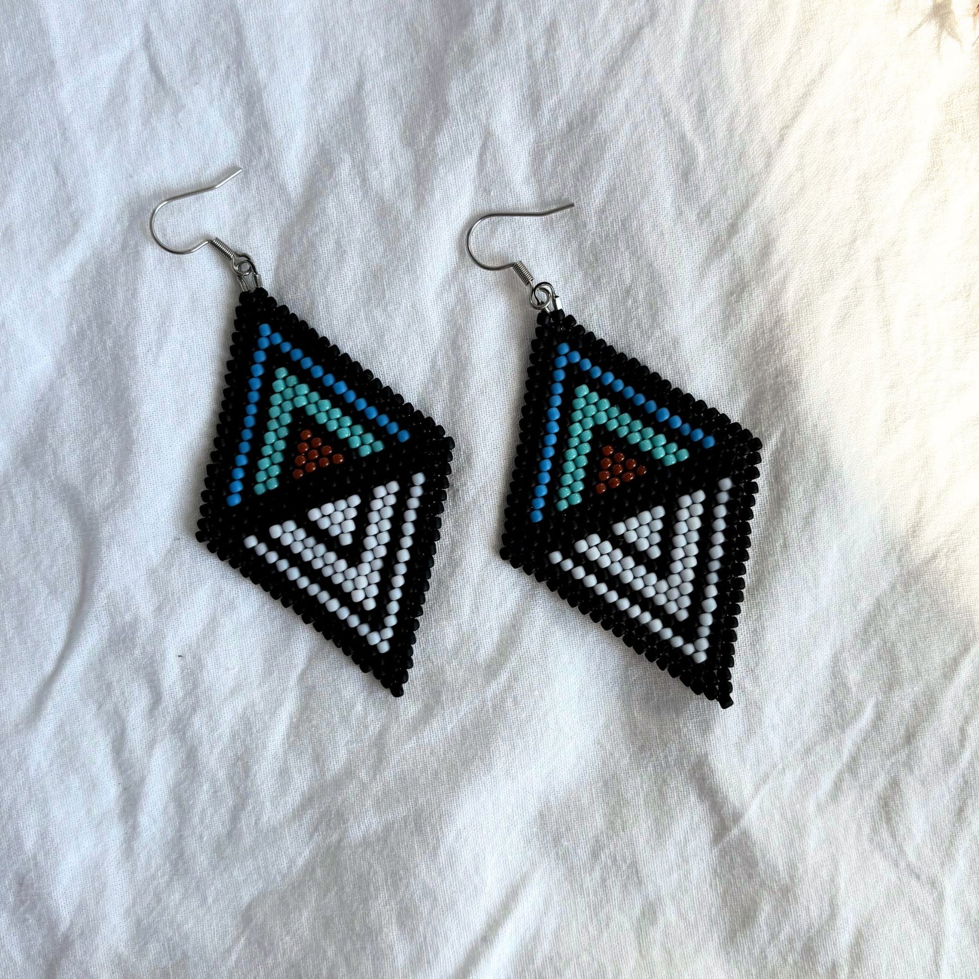Cercei handmade lungi realizați din mărgele delicate, cu design geometric simetric tip romb în culori negru, alb, albastru, turcoaz și portocaliu, cercei statement ușori, stil boho-elegant, bijuterii artizanale unicat potrivite pentru ținute de vară, evenimente sau cadou.